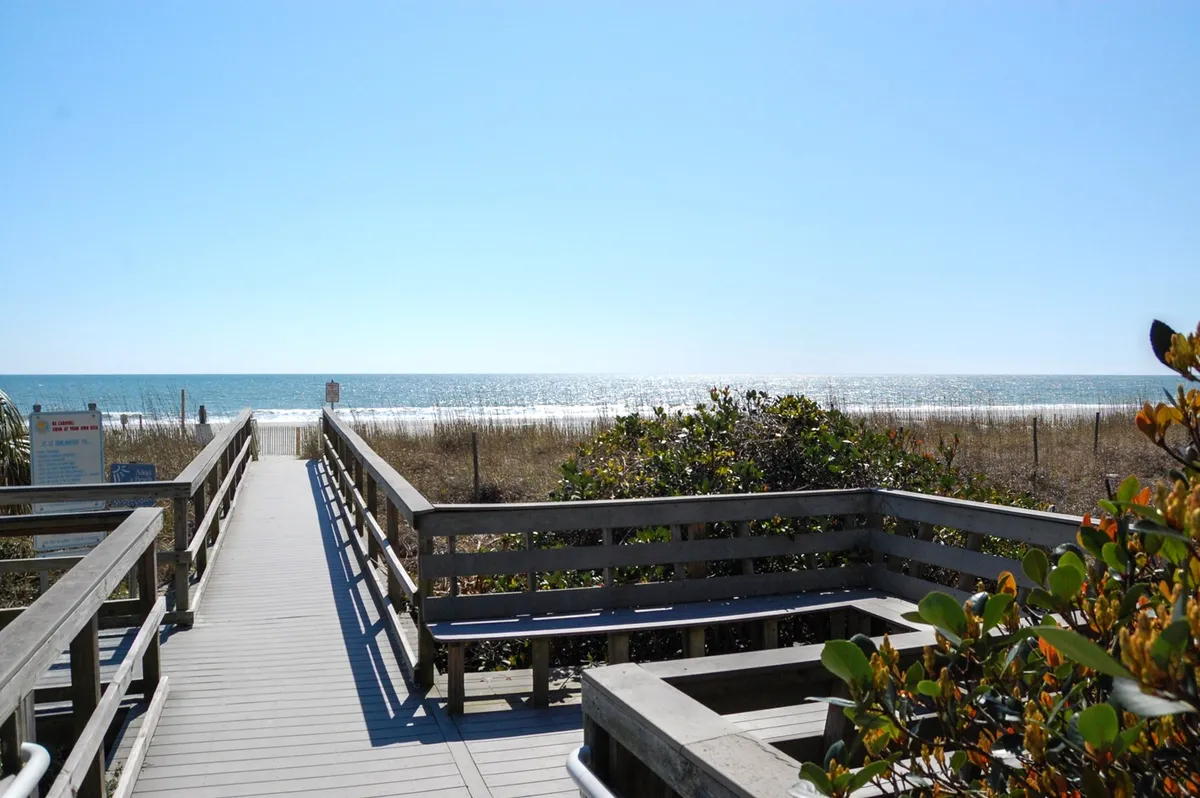 A wide wooden walkway leads over dune grass to the beach, with expansive ocean views stretching out under a clear sky. The scene is tranquil and inviting, showcasing the natural beauty and peaceful atmosphere of Myrtle Beach during quieter months.