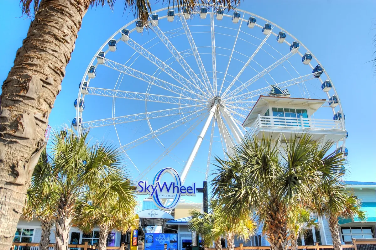 A festive scene featuring the iconic Myrtle Beach SkyWheel, framed by palm trees and an inviting entrance structure. People are visible, suggesting excitement and anticipation for the ride, with the SkyWheel towering above as a landmark attraction.