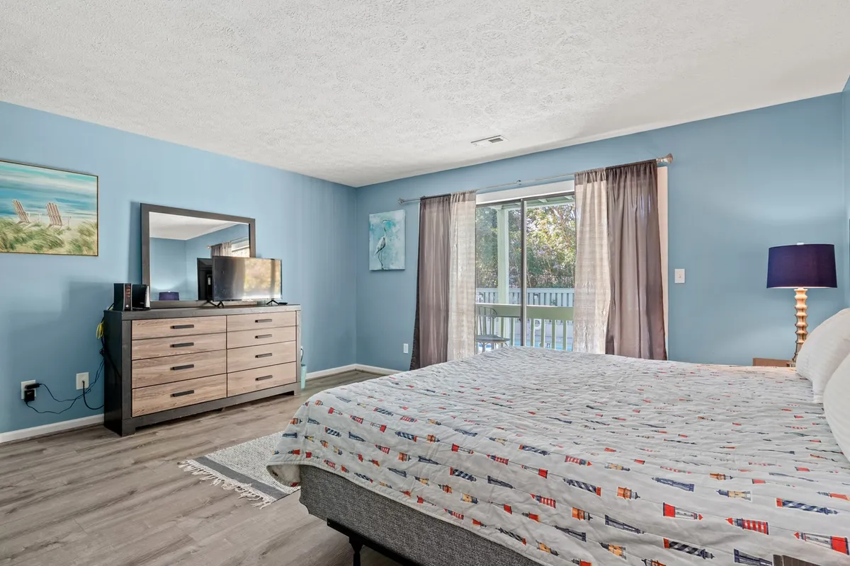 A spacious bedroom with a comfortable bed, dresser, and sliding glass door leading to a balcony. Artwork and soft furnishings add to the calming ambiance, and natural light fills the room, enhancing the sense of openness and relaxation.