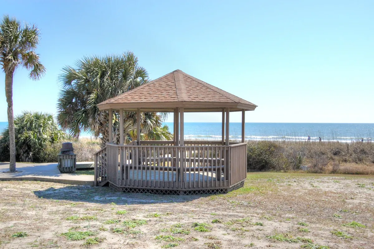A wooden gazebo sits by the beach, surrounded by palm trees and offering panoramic views of the ocean. The setting is calm, ideal for relaxing, watching sunsets, or enjoying the coastal breeze.