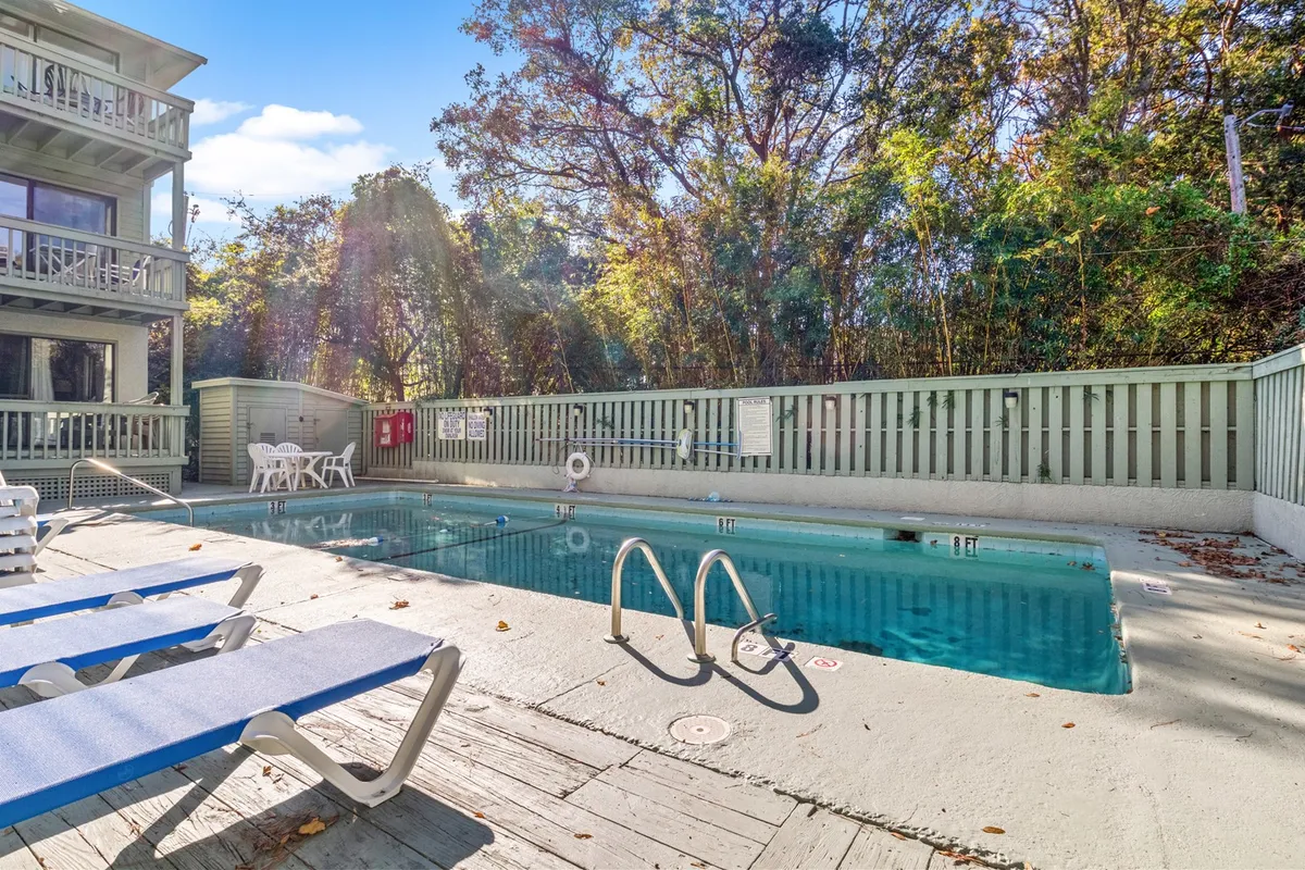 The image showcases the condo's sparkling swimming pool surrounded by inviting lounge chairs, lush trees, and the facade of the building, with an atmosphere perfect for relaxation and socializing. The scene evokes possibilities for sunset swims, afternoon BBQs, and peaceful mornings by the water, all just steps from the beach.