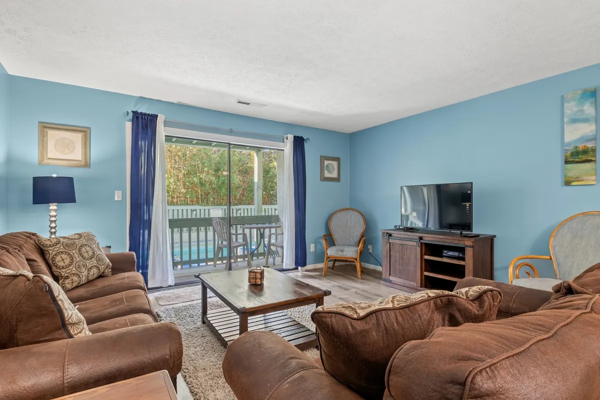 A spacious living room featuring comfortable brown sofas arranged in a welcoming layout, a wooden coffee table at the center, a flat-screen television set up for movie nights, and large glass sliding doors revealing serene pool views that infuse the space with natural light.