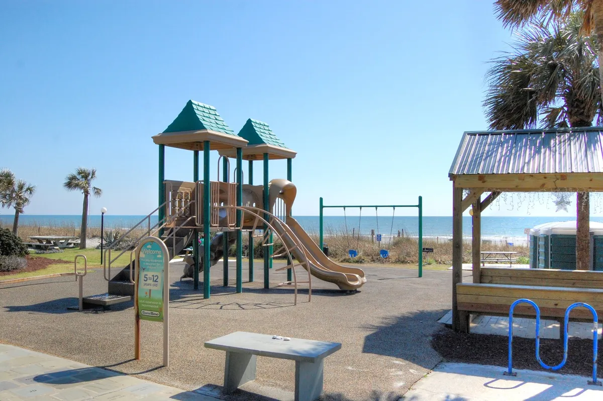 A lively beachfront playground scene with children playing on the swings and slides, families gathered at picnic tables, and an expansive ocean view as the backdrop. The mood is joyful and energetic, reflecting the fun and communal spirit of family vacations.