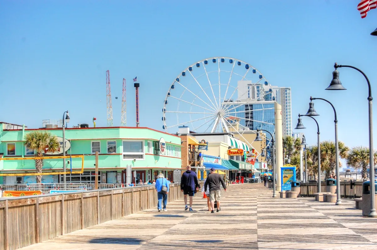 A lively Myrtle Beach boardwalk scene featuring the iconic SkyWheel, colorful storefronts, bustling crowds, and people enjoying rides and shopping. The atmosphere is festive and energetic, perfect for evenings out with friends.