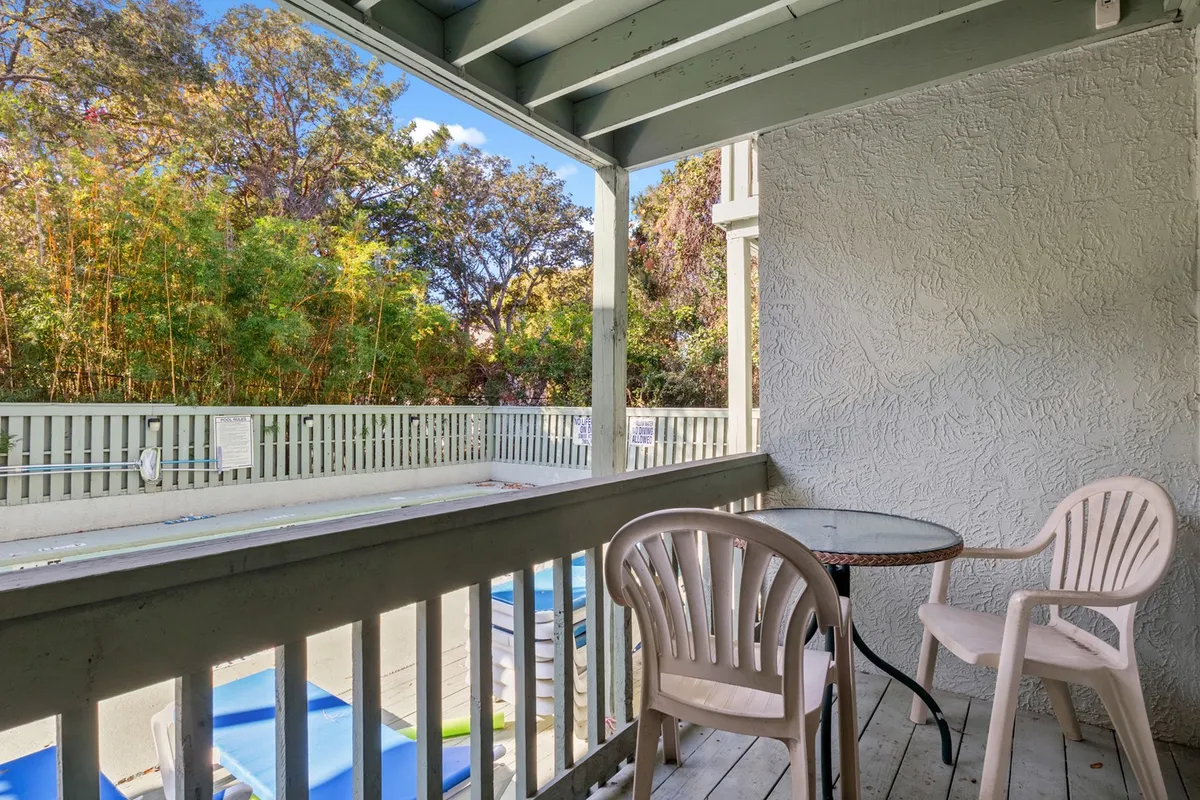 A small private balcony furnished with a round table and two plastic chairs, surrounded by lush greenery. The view from the balcony overlooks poolside areas, and the space is bathed in soft, natural light—ideal for enjoying coffee or reading a book outdoors.