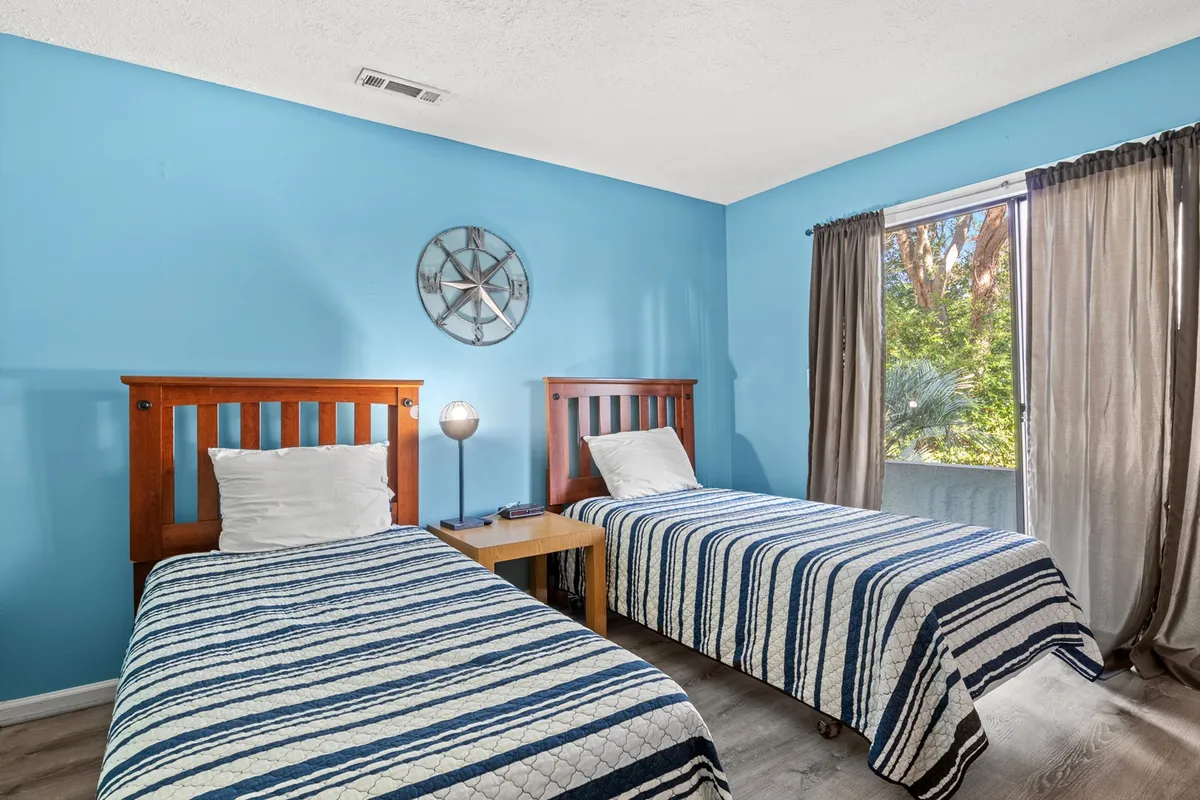 A cozy second bedroom with two twin beds, each covered in patterned bedding. Natural light streams in from a large window, and a decorative clock adds a touch of personality. The room is designed for comfort, making it ideal for children or friends sharing the space.
