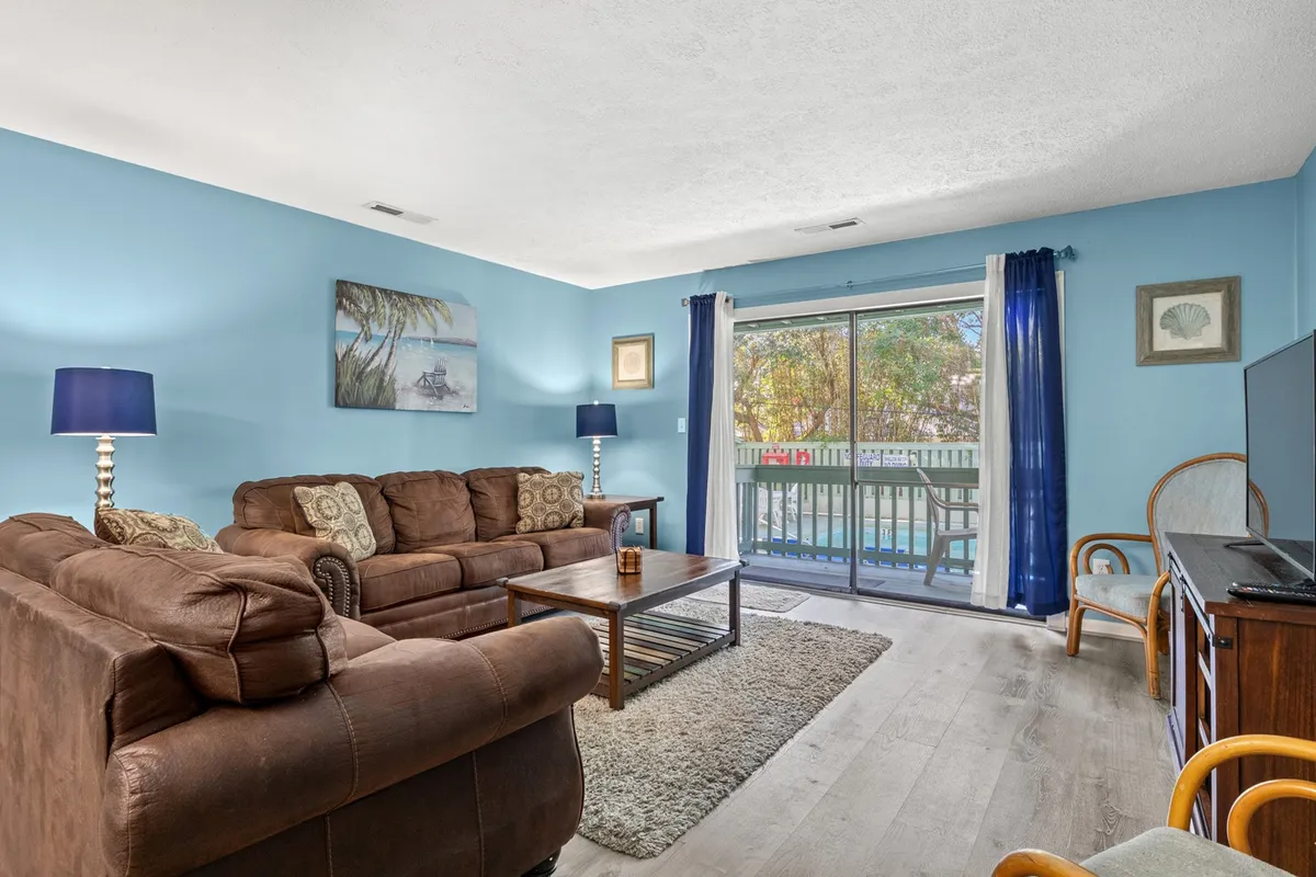 A comfortable brown leather sofa positioned to face a sliding glass door, with blue lamps and decorative wall art enhancing the homely, welcoming ambiance. The setup hints at intimate gatherings and relaxed entertainment, with pool views peeking through the door.